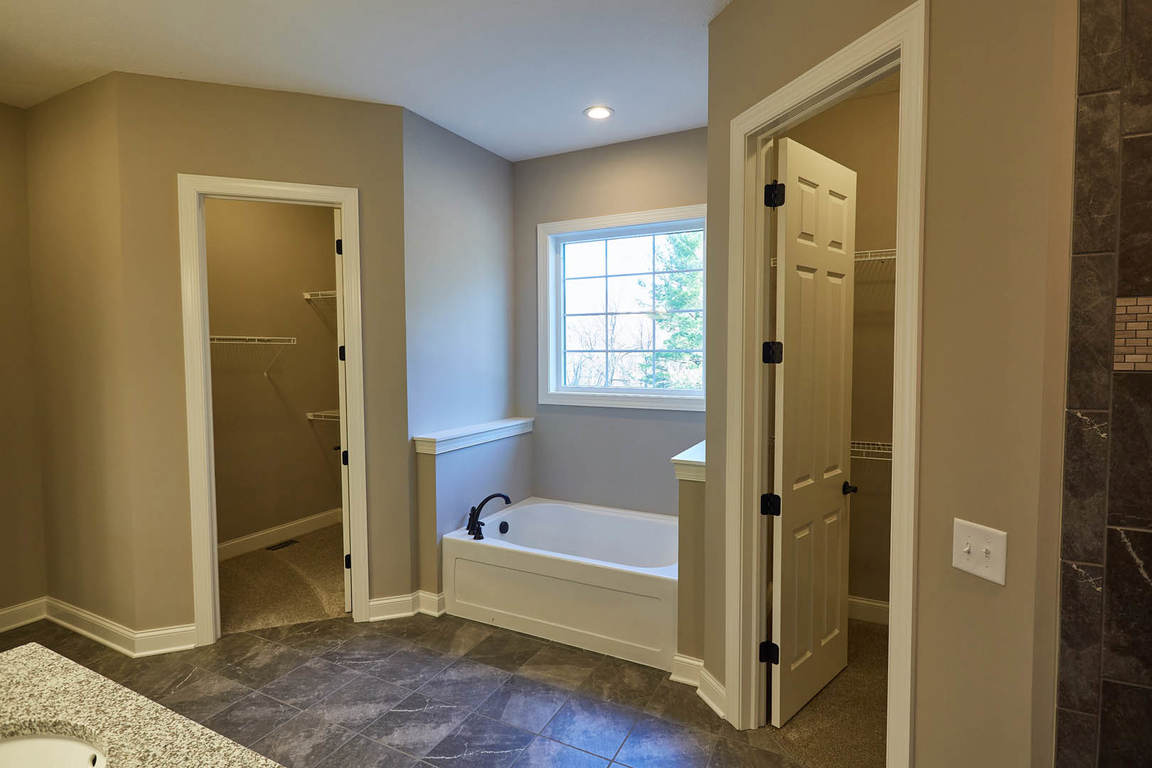 Bathroom with white bathtub featuring black faucets, glass-enclosed shower, grey tile flooring, wall-mounted light switch, window with multiple square panes, and closet with