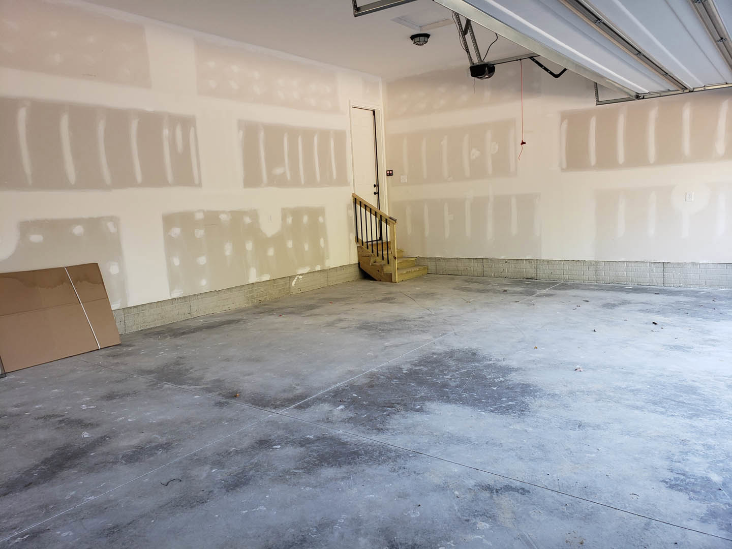 Concrete floor with a wooden staircase featuring black railings, white plaster walls, and a cardboard box near the base of the stairs in an empty room.