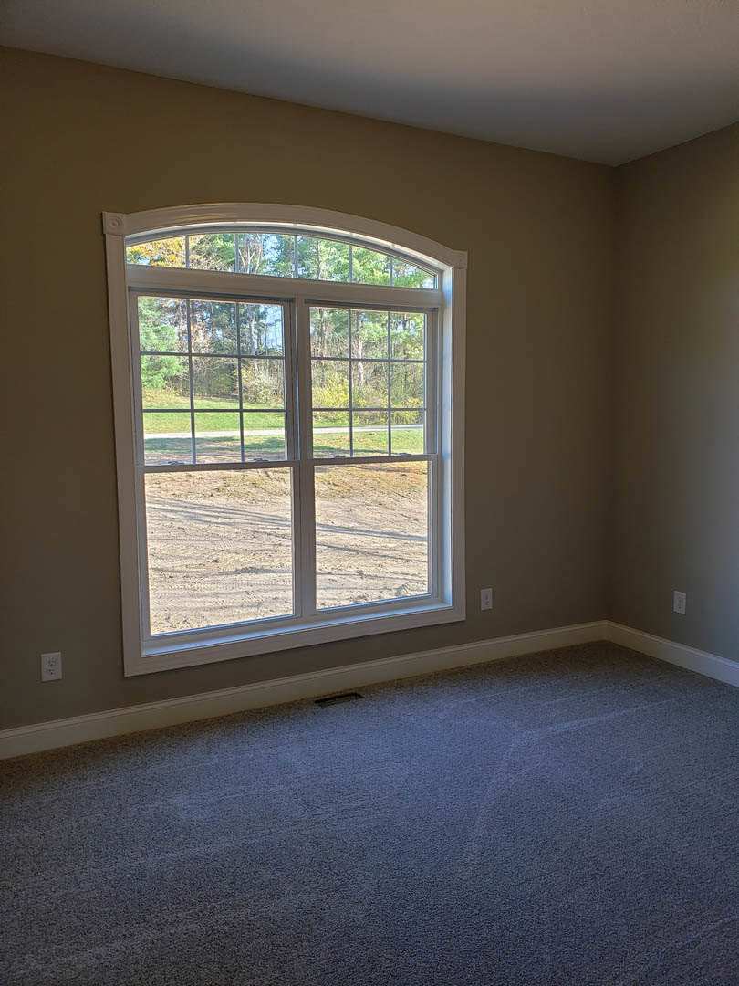 Carpeted room with white walls, brown trim, and large window overlooking dirt field, grass, and trees