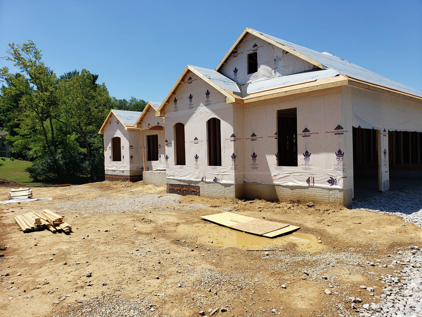 Framed house under construction with exposed wood, surrounded by trees and grass, unfinished sidewalk and door visible