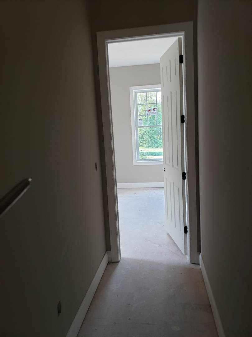 White hallway with open door, sunlight streaming onto pale flooring, white plaster walls, window with sign visible, modern door handle.