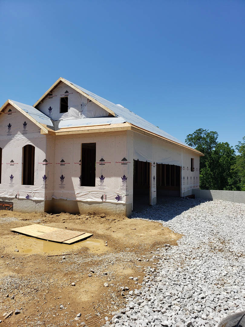 Modern home under construction with exposed framing, gravel driveway, plastic sheeting covering exterior walls, and clear blue sky overhead