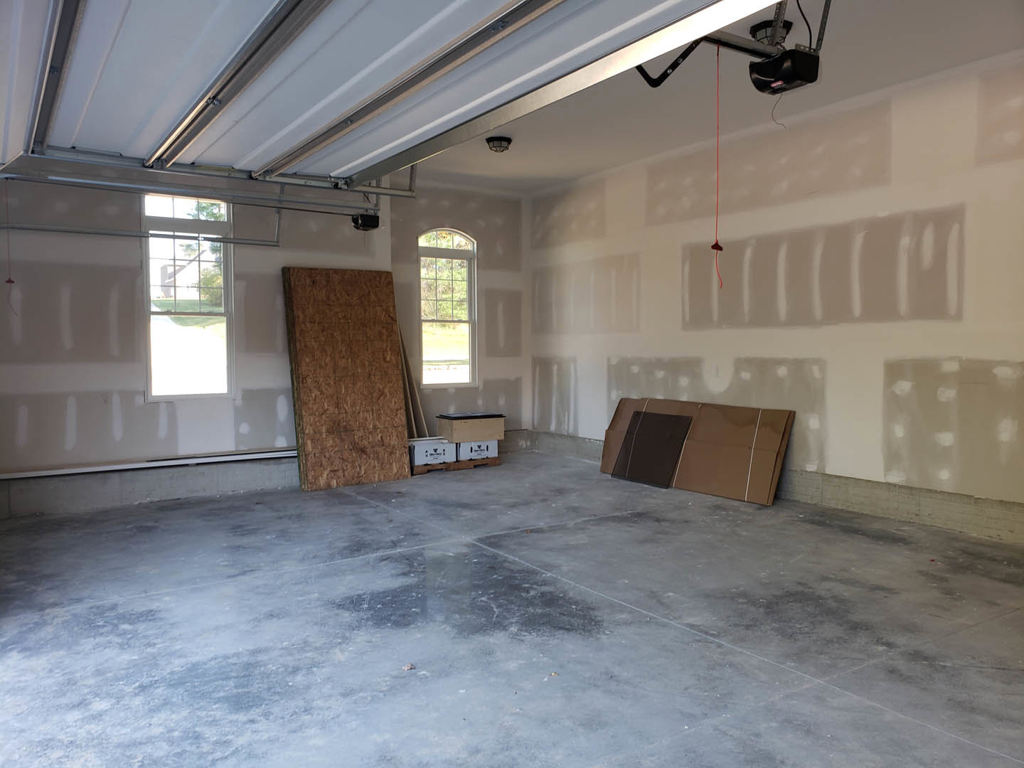 Unfinished room with plaster walls, plywood surfaces, concrete floor scattered with boxes, garage door, and window showing outdoor view