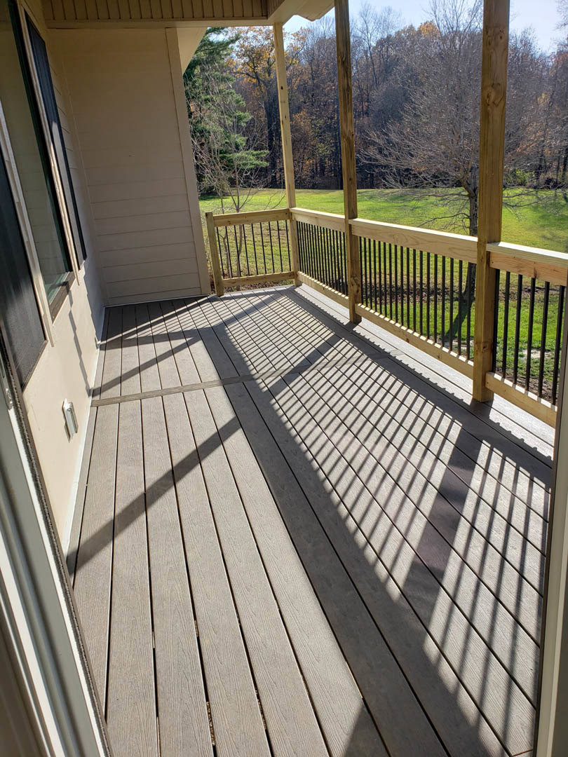 Wooden deck with black metal railing, white exterior wall with black-framed window, grassy yard and fence in the background