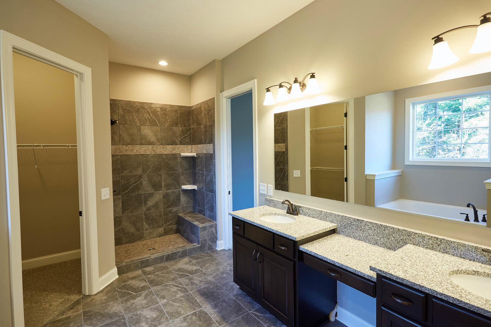 Bathroom featuring a large wall mirror above a granite countertop, glass-enclosed shower with tile walls, white cabinetry, and a window overlooking trees.