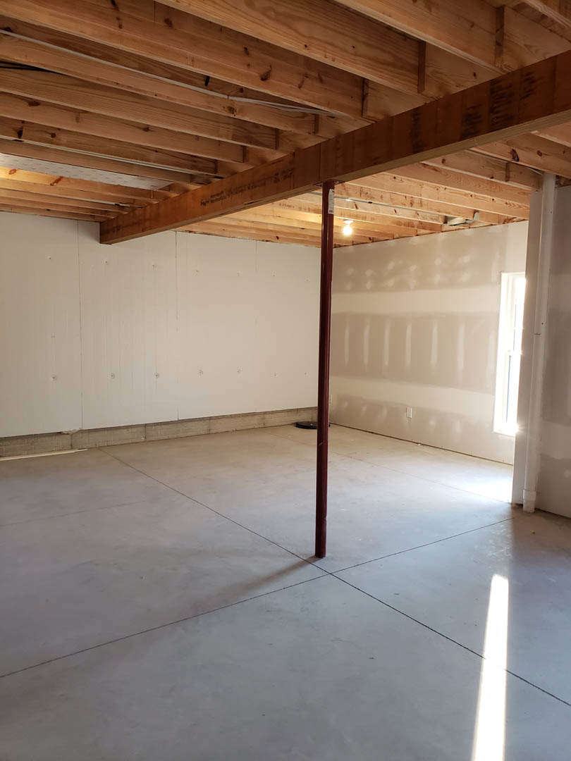 Concrete floor with a central metal pole, white plaster walls, large window letting in natural light, exposed wooden ceiling beams