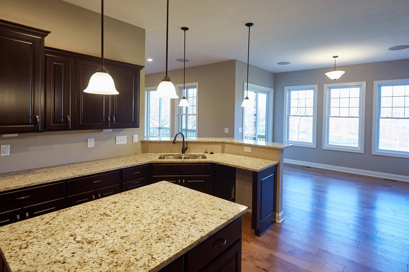 Kitchen with brown speckled granite countertops, hardwood flooring reflecting blue light, white-framed window, wood cabinetry, stainless steel sink, and modern light fixture.