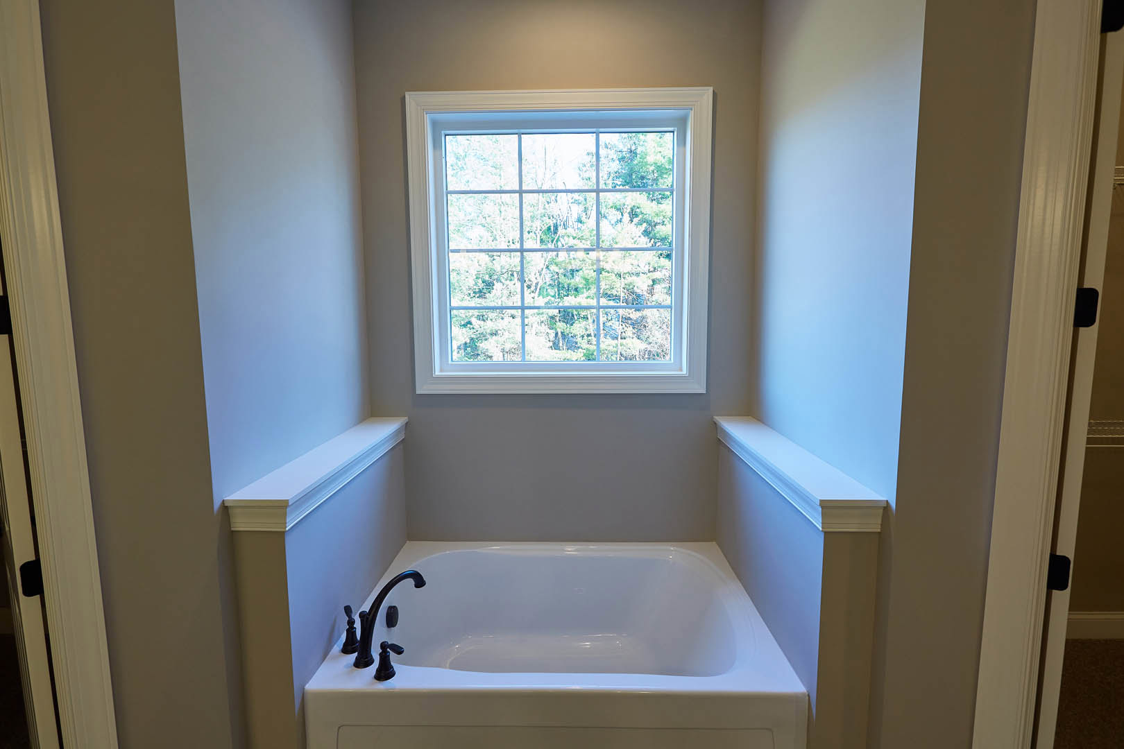 Freestanding white bathtub with chrome faucet beneath large window overlooking trees, white shelf mounted on wall, light-colored surfaces throughout bathroom