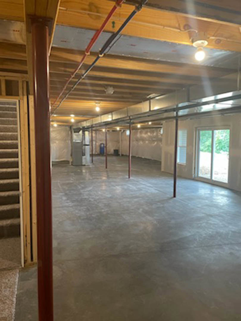 Concrete floor with red support pole, white-framed window, glass door, exposed ceiling pipes, brown and white walls, staircase leading to upper level