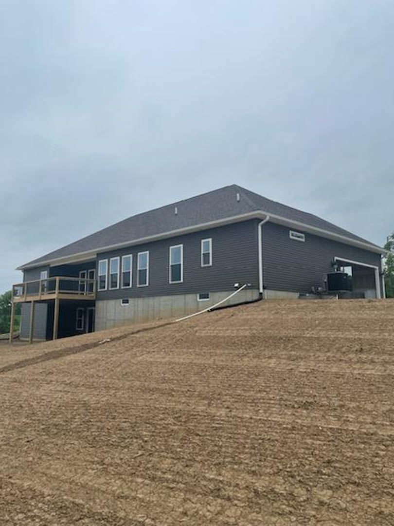 Two-story house with grey roof and balcony, situated on a grassy hill, white exterior walls, large windows, garden hose coiled on the side, cloudy sky overhead