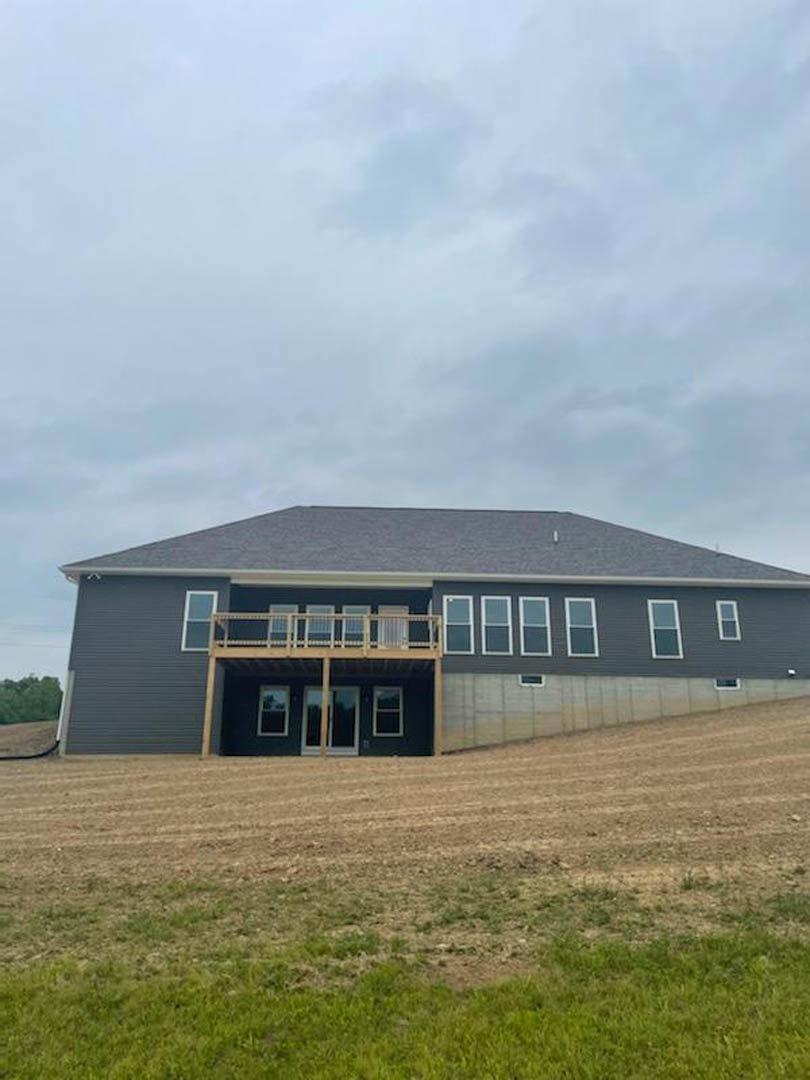 Two-story house with wide wooden deck overlooking expansive grassy field, large windows, and light-colored siding under partly cloudy sky