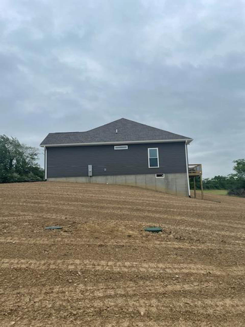Modern home with gray roof and white siding sits atop a grassy hill, surrounded by a dirt field with tire tracks and a wooden fence; scattered trees and clouds in the sky complete