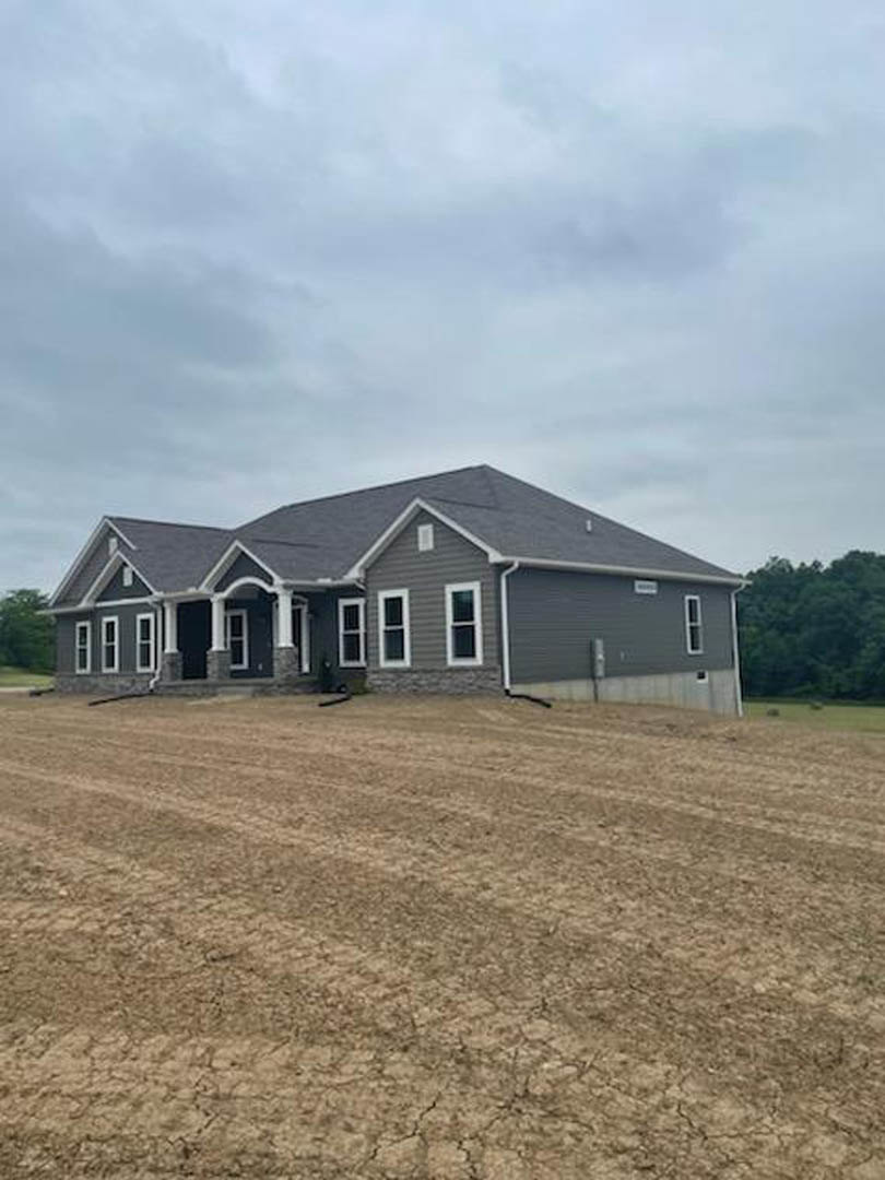 Grey-roofed house with white siding and multiple windows, surrounded by a dirt field and sparse grass, cloudy sky overhead, distant trees and fence visible in background