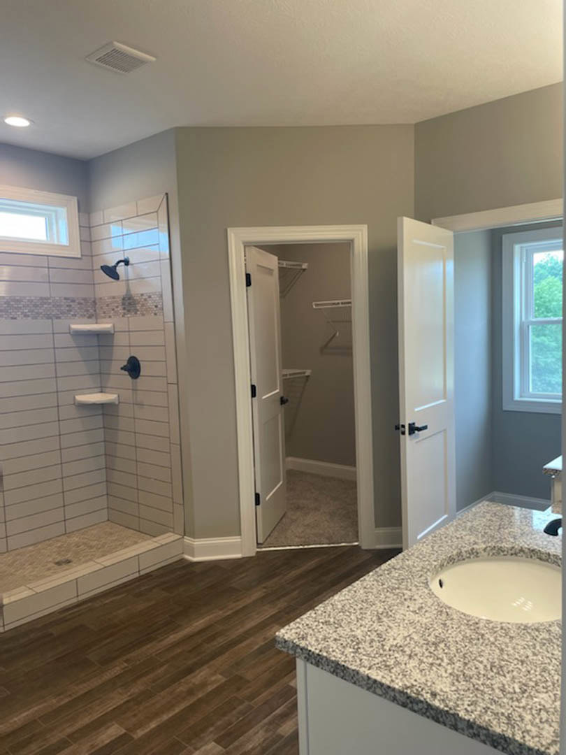 Bathroom with wood flooring, marble countertop, white vessel sink, black speckled surface, glass shower enclosure, and window overlooking green trees.