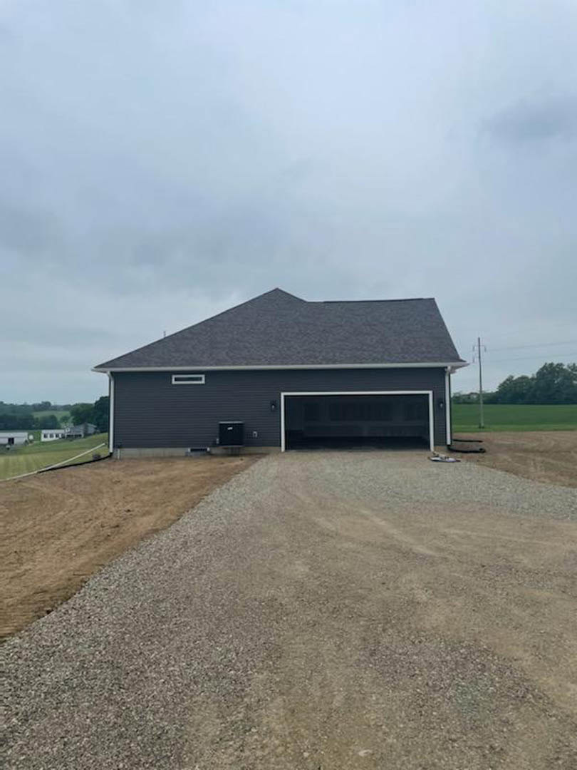 Two-story house with attached garage, white siding, gray shingle roof, gravel driveway, grassy yard, scattered trees in background, partly cloudy sky