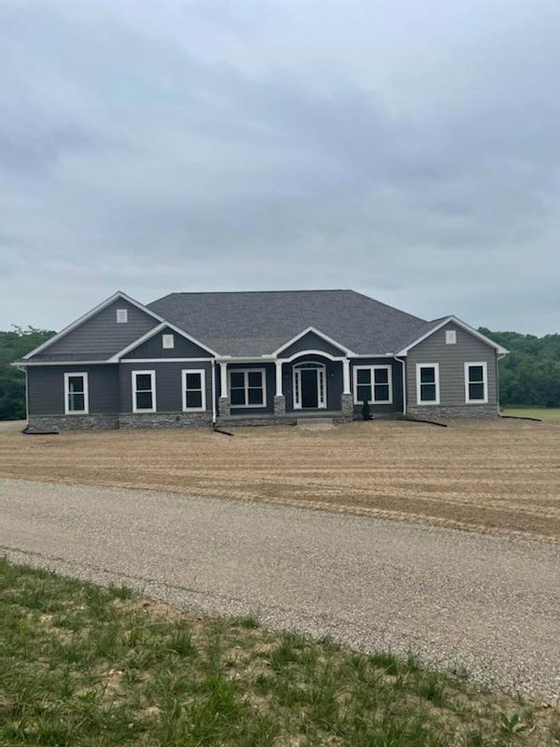 Two-story house with grey roof, white-framed windows and door, dirt road and patch of bare earth in front, surrounded by grass under cloudy sky