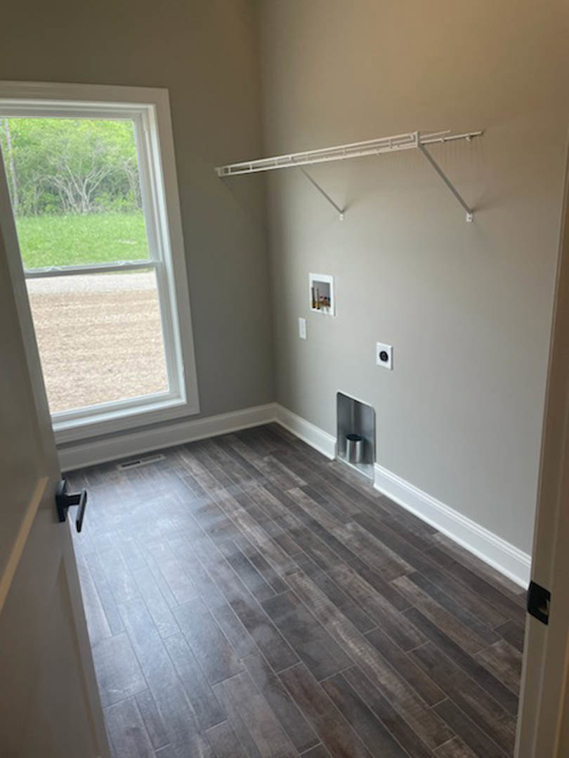 Wood flooring in a room with a large window overlooking a grassy field, white plaster walls, metal cylinder fixture, white pipe, and a trash can near the wall.