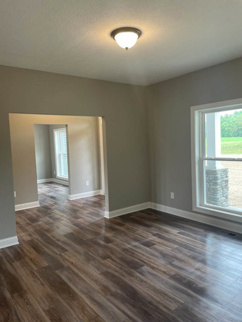 Open-concept room with hardwood flooring, modern ceiling light fixture, white brick accent wall, large window framed by a stone pillar, and an open doorway.