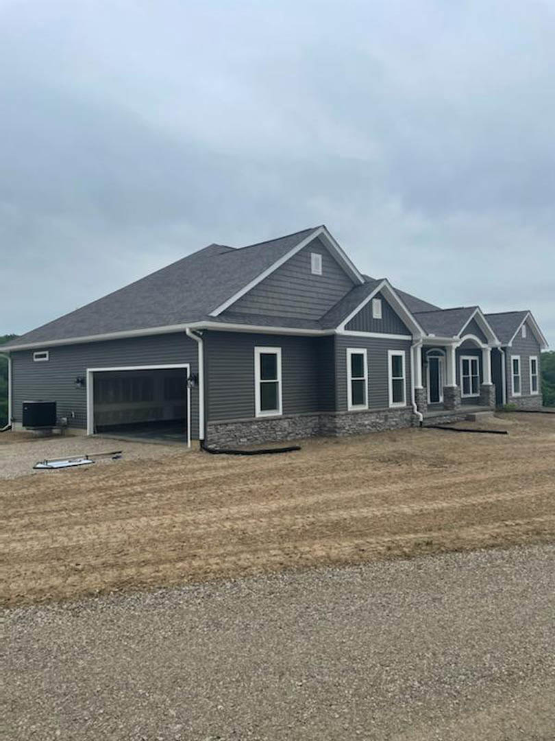 Two-story house with gray roof, attached garage featuring a black garage door, light siding, large windows, and dirt landscaping in front under a partly cloudy sky.