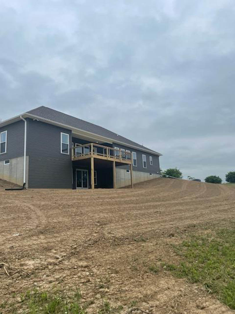 Modern house with wood deck overlooking expansive dirt field, cloudy sky above, sparse grass and trees at property edge