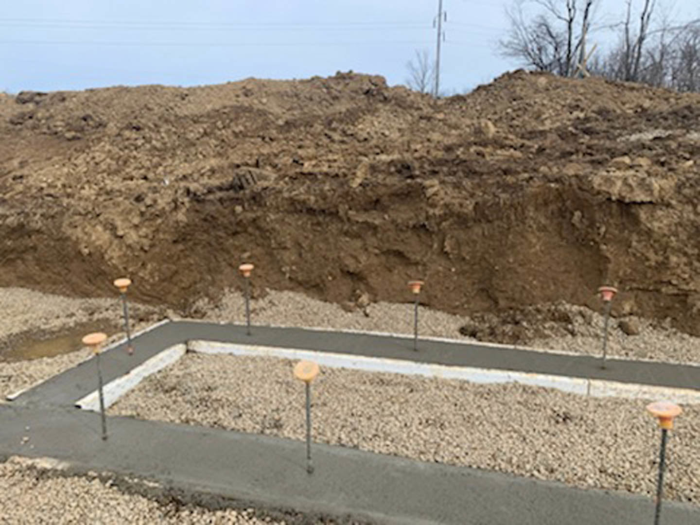 Concrete foundation with vertical poles set in bare dirt, surrounded by soil and sparse vegetation, under an open sky.