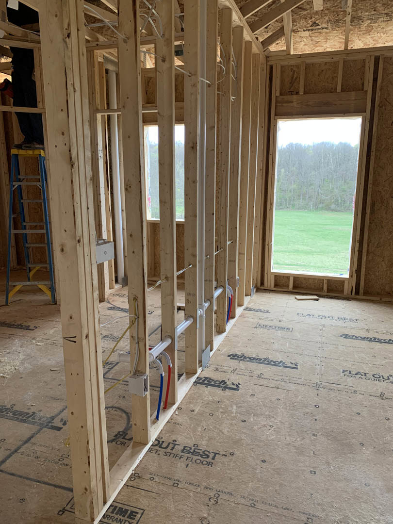 Exposed wood beams and framing with visible pipes and wires, ladder leaning against unfinished wall, large window overlooking distant forest trees