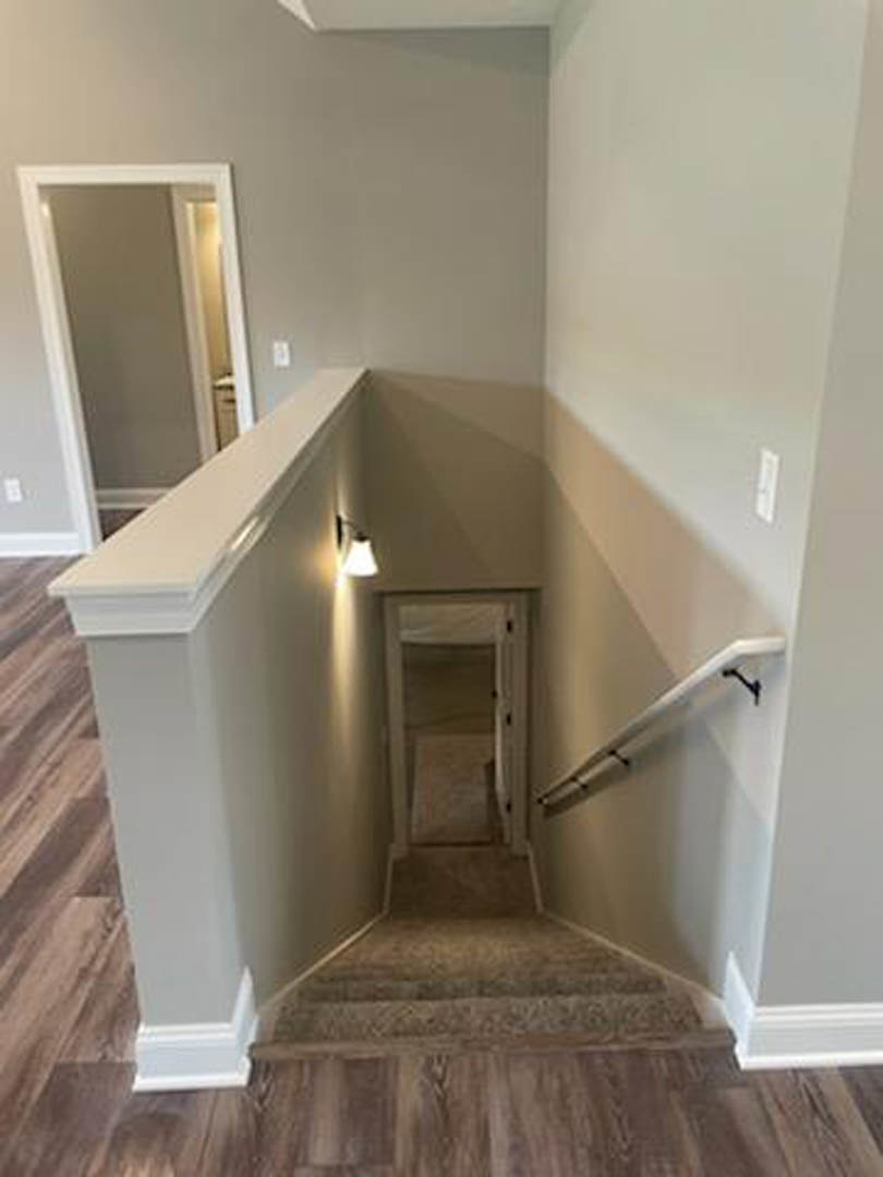 Wood staircase with metal handrail ascending to a white door set in a grey wall, laminate flooring and plaster walls, interior lighting fixture visible.