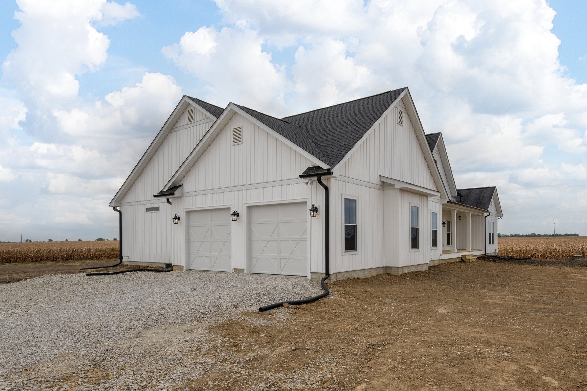 White house with black roof, three garages featuring white doors with X design, gravel driveway, black outdoor lights above garage doors, black pipe on ground near garage, cloudy