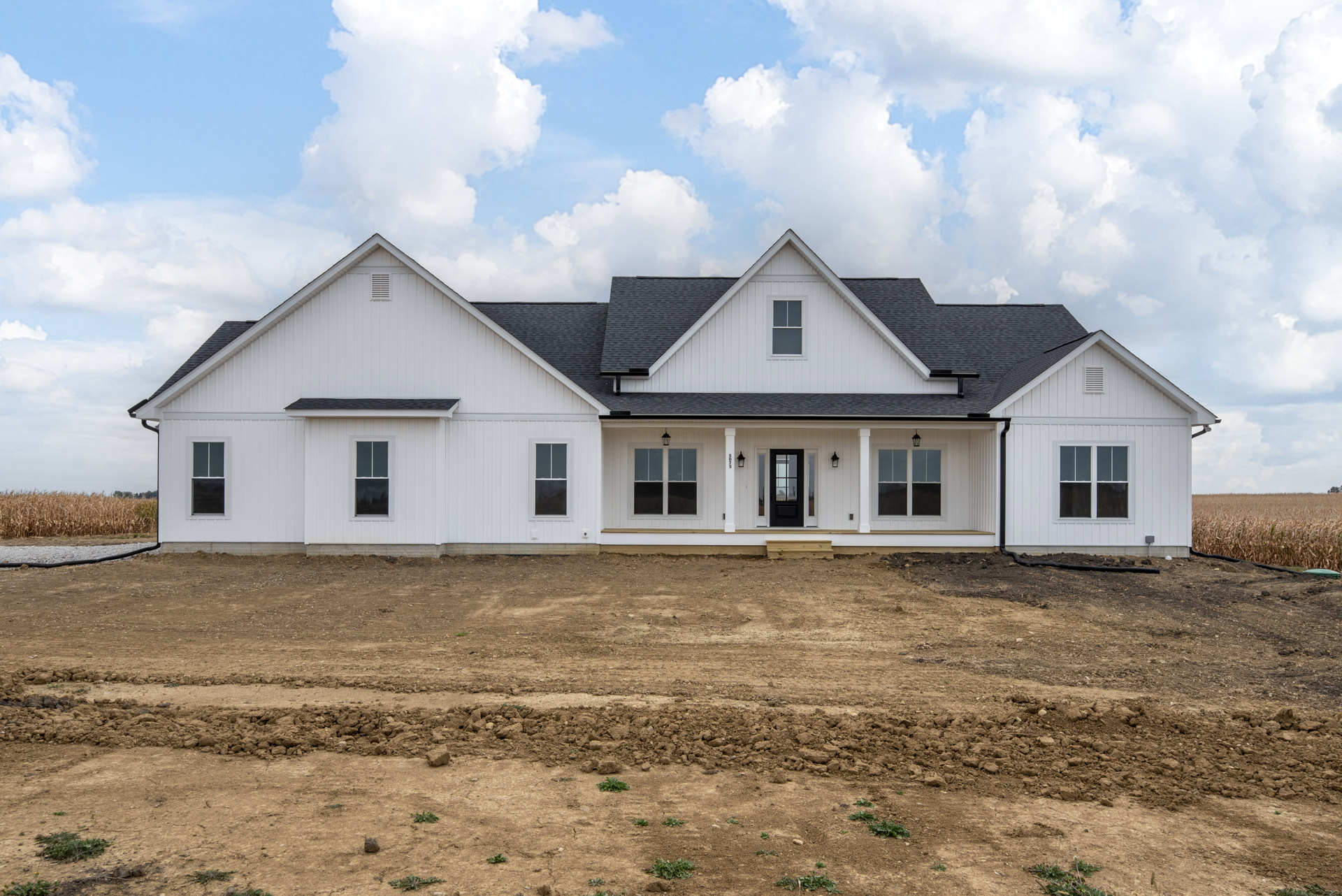 White house with black roof and black door, white-framed windows, dirt field in foreground with pile of soil, cloudy sky above