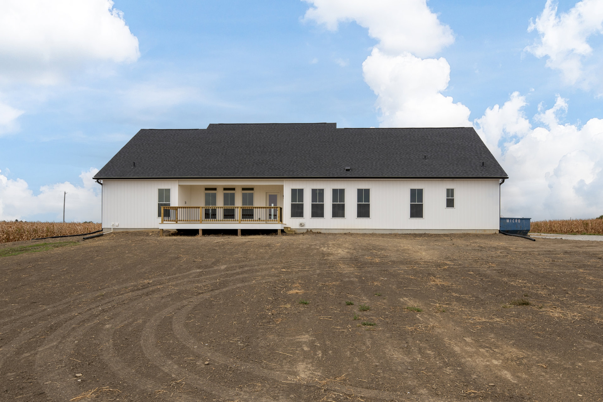 White modern house with flat black roof, large windows, covered porch, upper balcony with metal railing, surrounded by dirt lot with patches of grass.