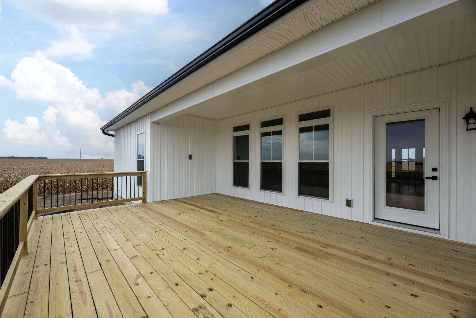 White siding house with large wooden deck, black railing, glass door, and white-framed windows overlooking corn field under partly cloudy sky