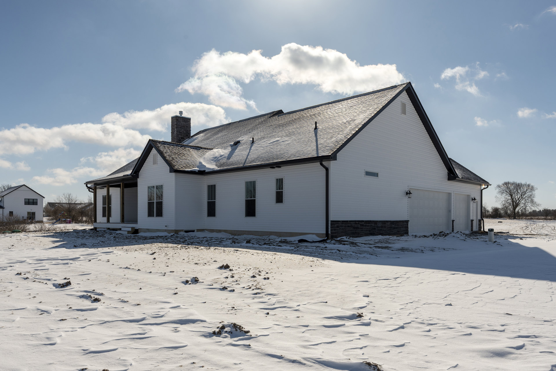 White house with black roof and brick chimney topped with a cross, snow covering roof and ground, visible tracks in snow, cloudy winter sky
