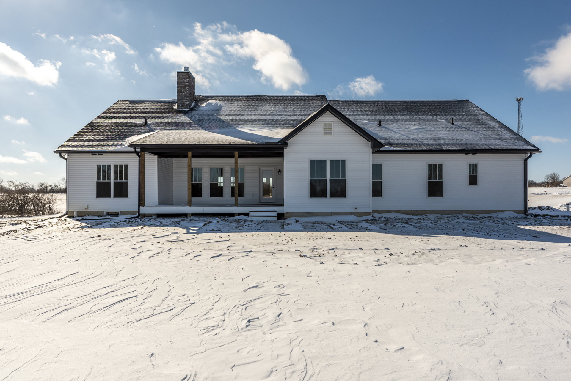 Two-story home with white siding, black front door, white-framed windows, and brick chimney, snow covering roof and ground, cloudy winter sky in background