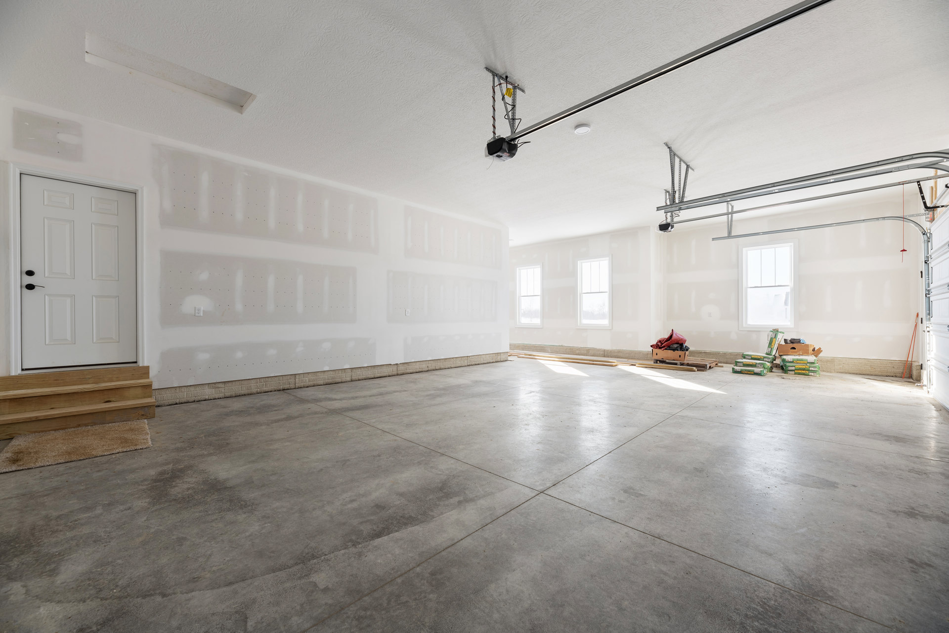 Concrete floor room with white plaster walls, large window with white frame, white door featuring black handles, and a box filled with plastic bags near the wall.