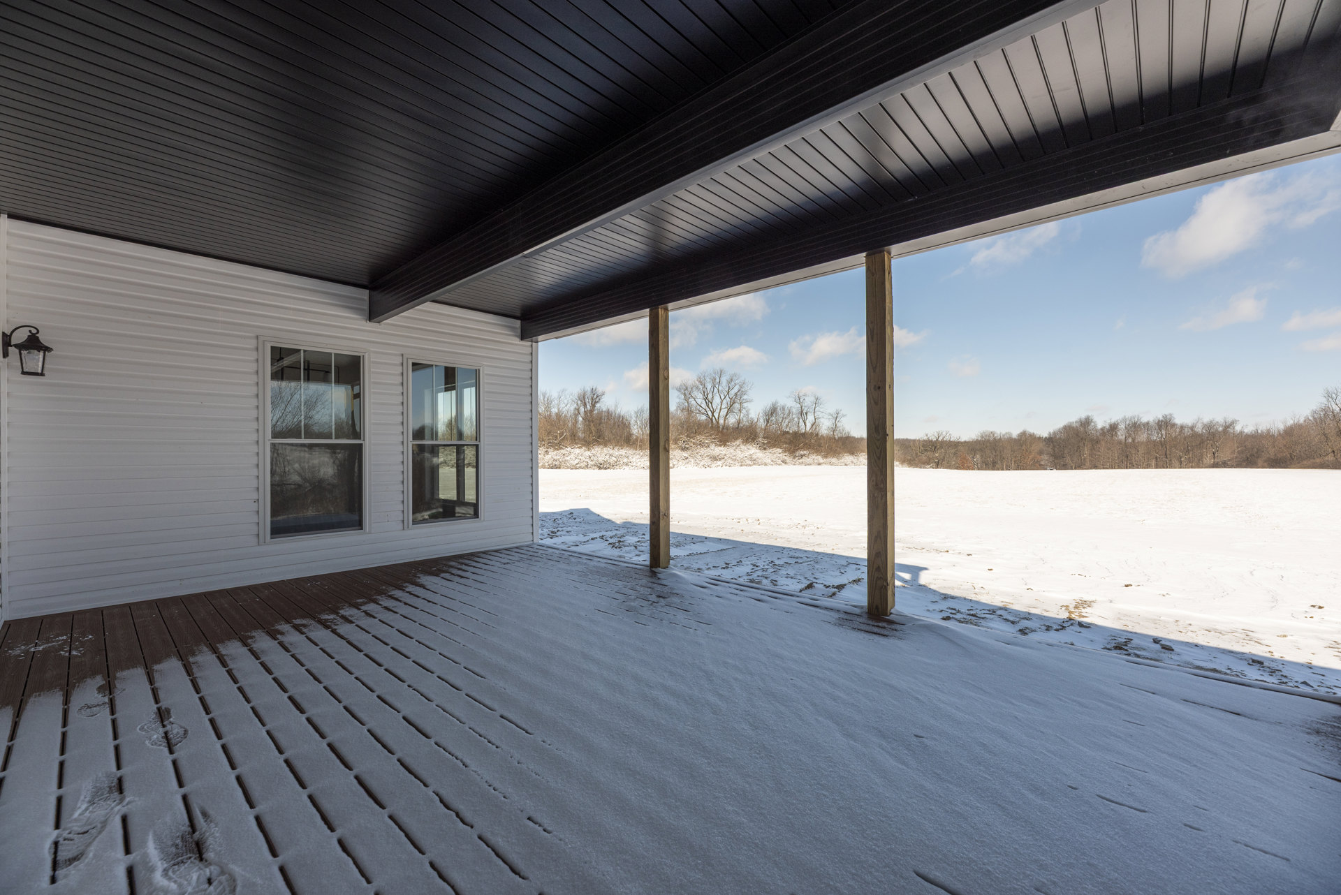 Covered front porch with snow on the ground, wooden support pole, windows reflecting trees, and a close-up of an exterior lamp.