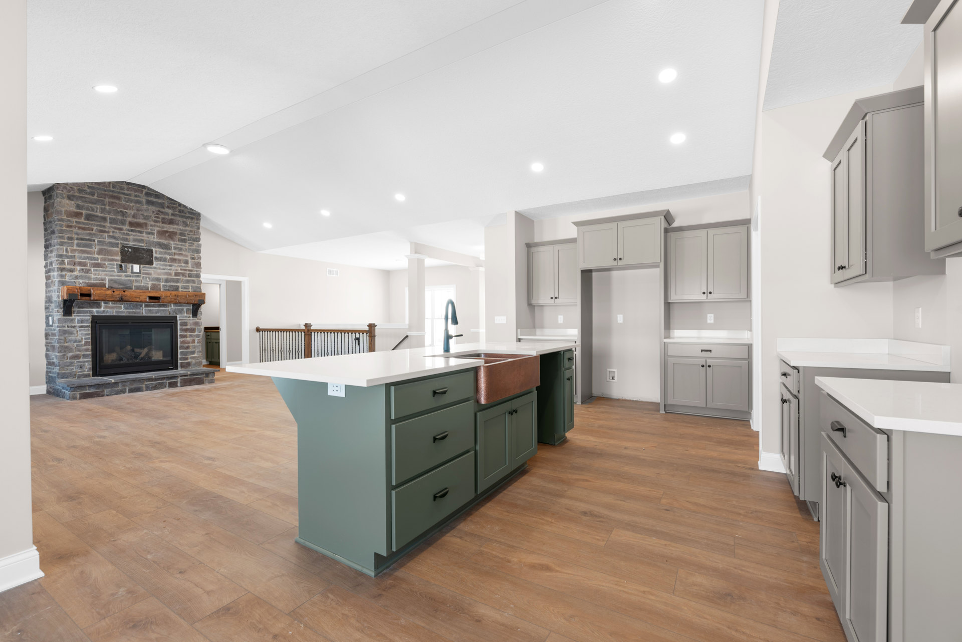 Spacious kitchen featuring a wood-burning fireplace with stacked logs, copper sink set in a central island, white cabinetry with black handles, wood flooring, and a green