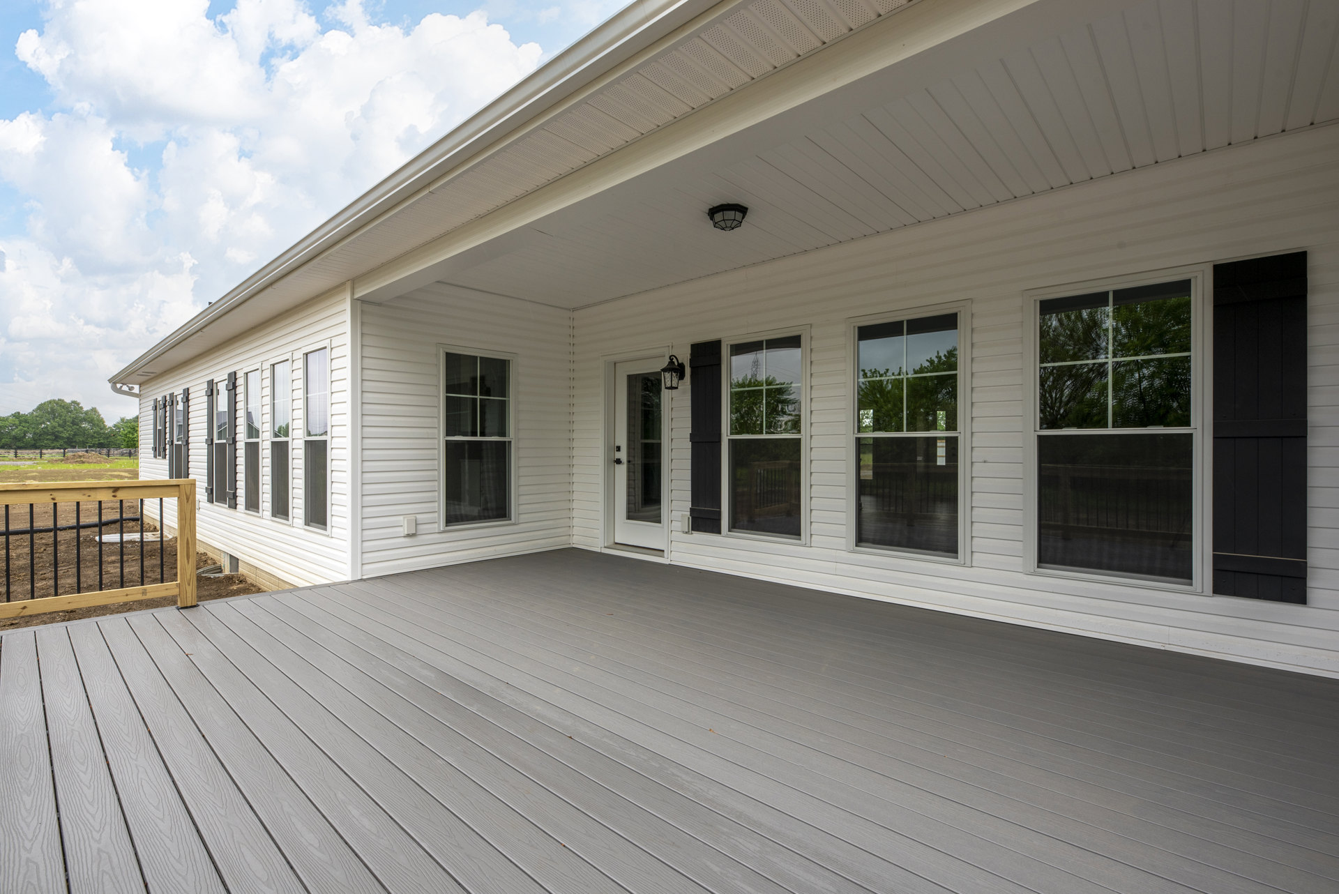 White siding exterior with wooden deck and railing, multiple windows reflecting nearby trees, under partly cloudy sky