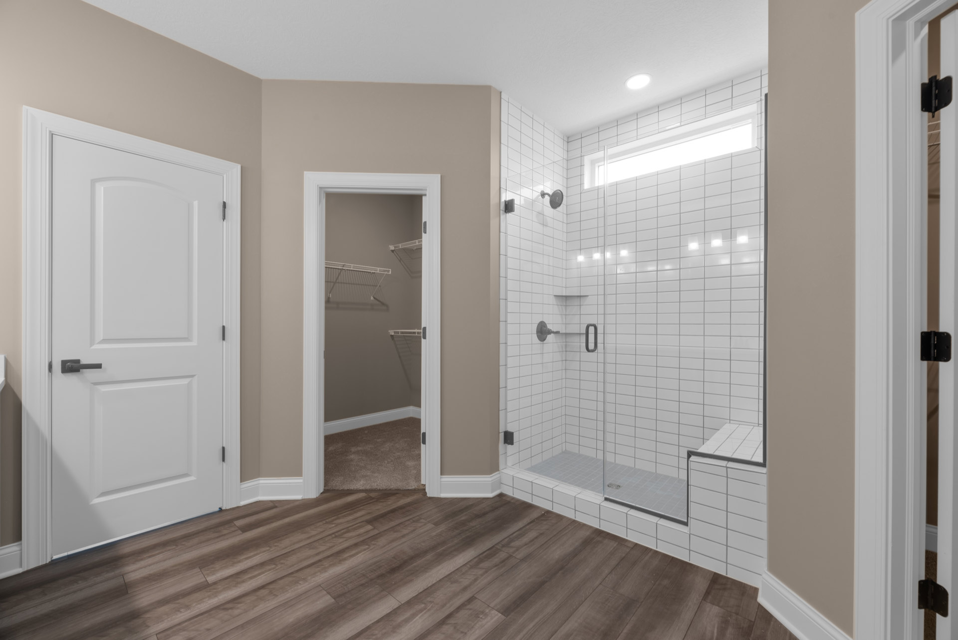 Bathroom with glass shower enclosure, white door with silver handle and black hinge, wood laminate flooring partially covered by beige carpet, built-in closet with white shelves