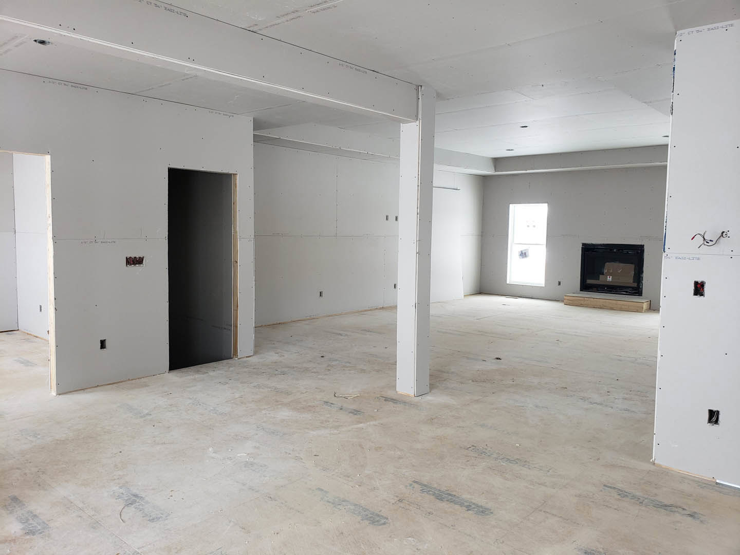 Modern living room featuring a black fireplace with glass doors, white paneled walls, large window displaying framed pictures, white flooring, and structural pillars.