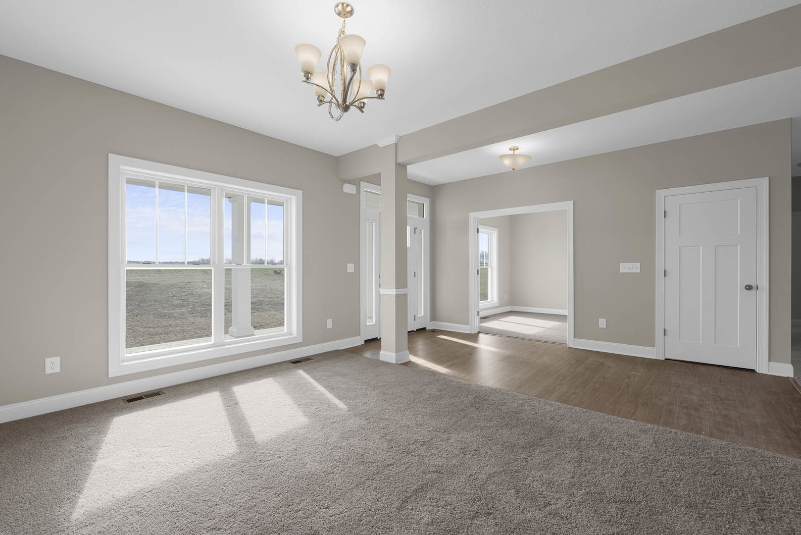 Chandelier hanging from white ceiling, large windows with field view, white door with black knobs, carpeted floor, light gray walls with crown molding