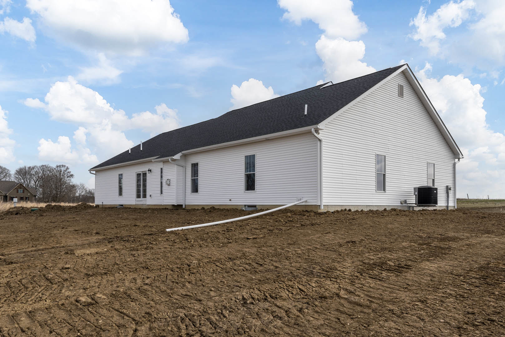 White house with black roof beside large dirt field, white pipe lying on ground, sparse grass, cloudy sky overhead, rural landscape.