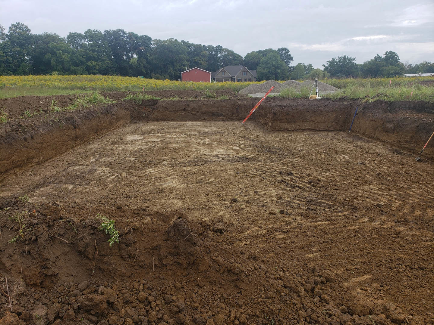 Dirt lot with excavated hole, red and white pole, red barn with white roof, grey-roofed house, cluster of distant trees under partly cloudy sky