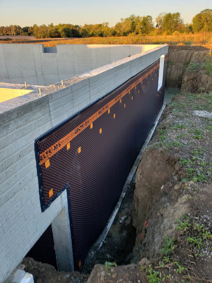 Concrete foundation with black and orange tape, dirt excavation in foreground, grassy area and trees in background under clear sky