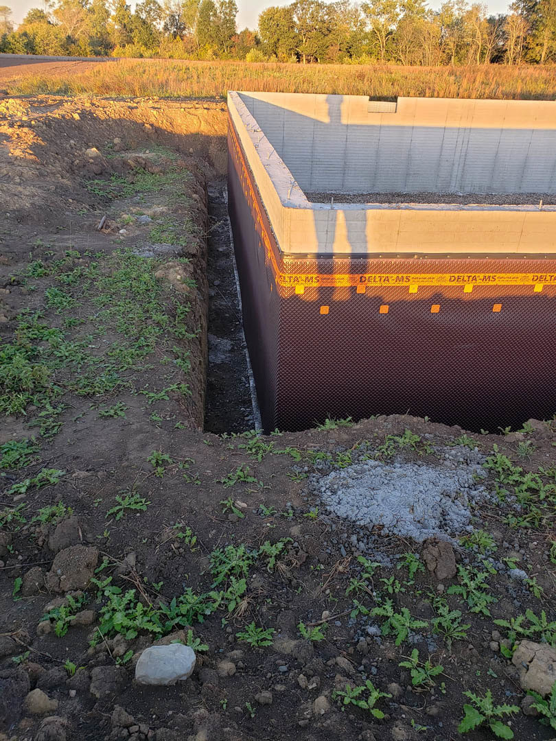 Partially constructed home with exposed foundation, white exterior wall, surrounding dirt and rocks, scattered trees in background, open sky above