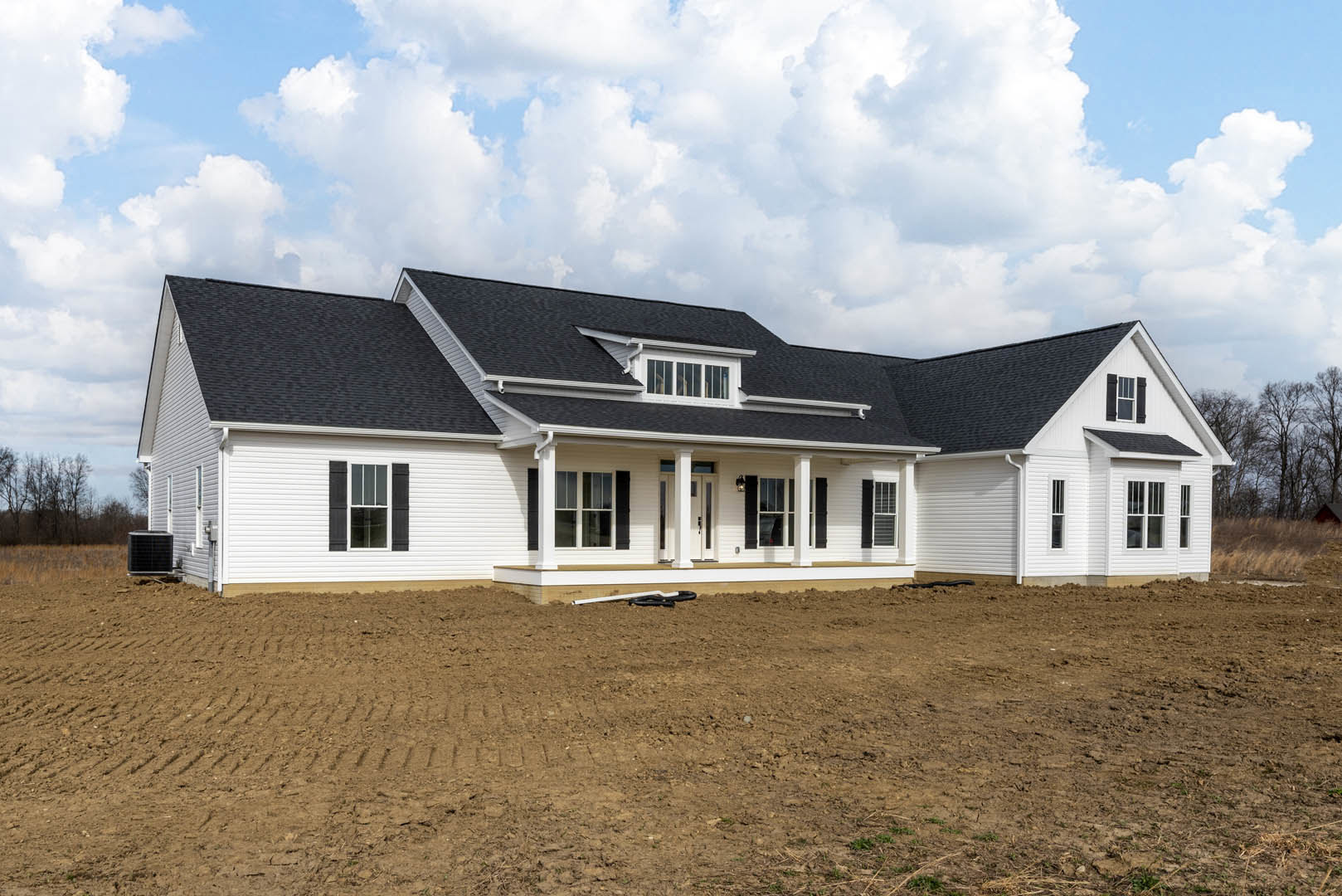 Two-story house with black roof and black shuttered window, surrounded by dirt field with garden hose and black grate object on ground, under cloudy sky.