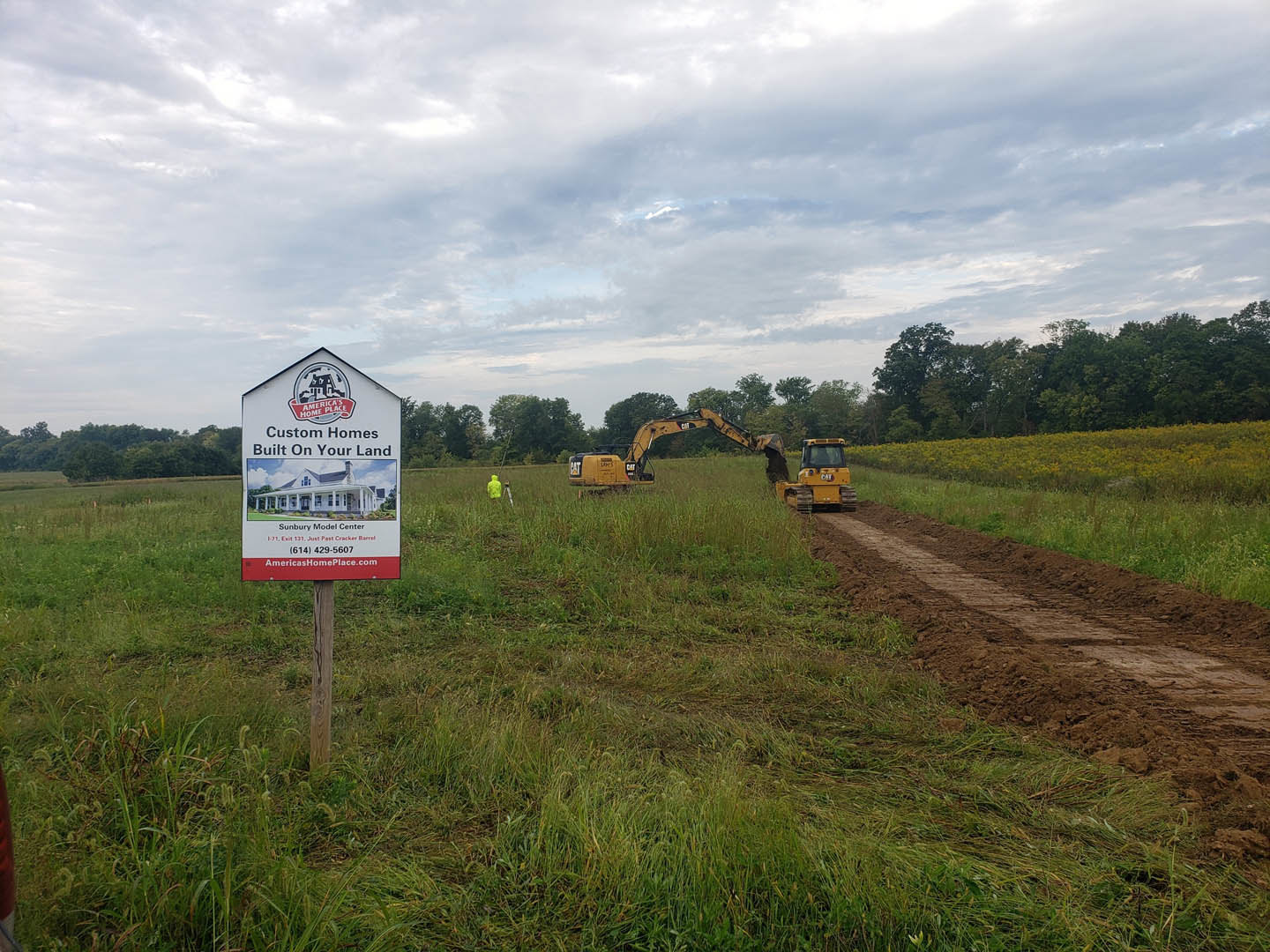 Bulldozers and construction equipment on grassy land lot, site sign in foreground, trees and cloudy sky in background