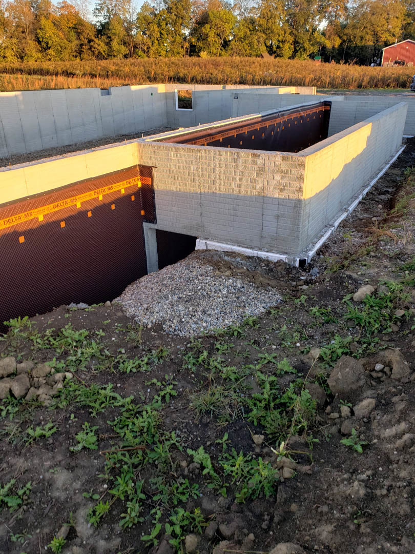 Partially built custom home with exposed concrete walls, black waterproofing strip, and large gravel pile in foreground; grassy field and trees in background.
