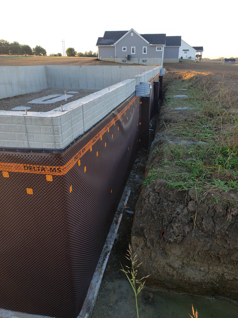 Modern home with light-colored siding, large windows, and sloped roof, landscaped front yard with green grass and small plants, second house visible in the background under clear