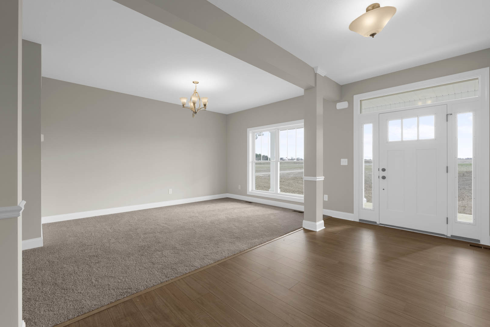 White paneled door and large window overlooking a grassy field, beige carpet flooring, white walls with crown molding, close-up of a modern chandelier hanging from plaster ceiling