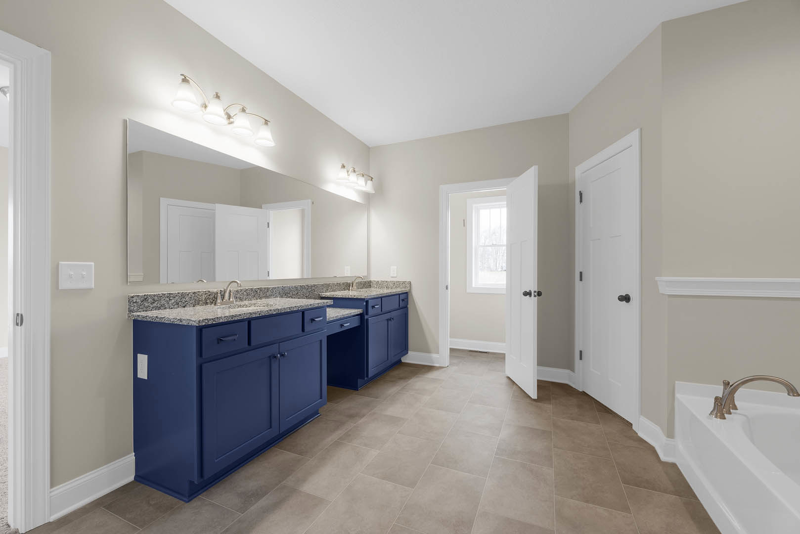 Blue bathroom vanity with marble countertop, chrome faucet, row of white lights above mirror, white door with black knob, white light switch on wall, tile flooring.